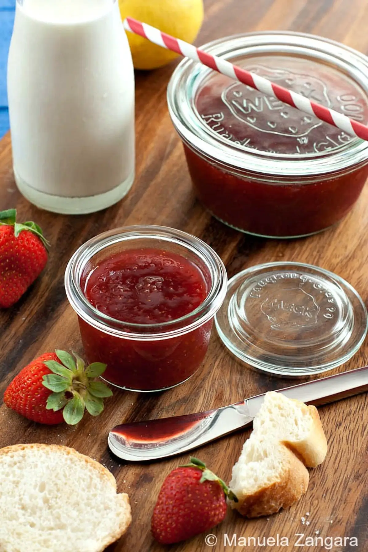 Glass jar filled with strawberry preserve served with fresh bread and whole strawberries on a wooden table.