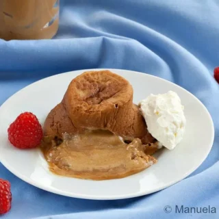 A warm molten dulce de leche cake on a white plate with whipped cream and raspberries on the side.