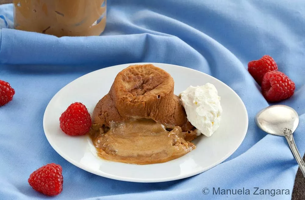 A warm molten dulce de leche cake on a white plate with whipped cream and raspberries on the side.