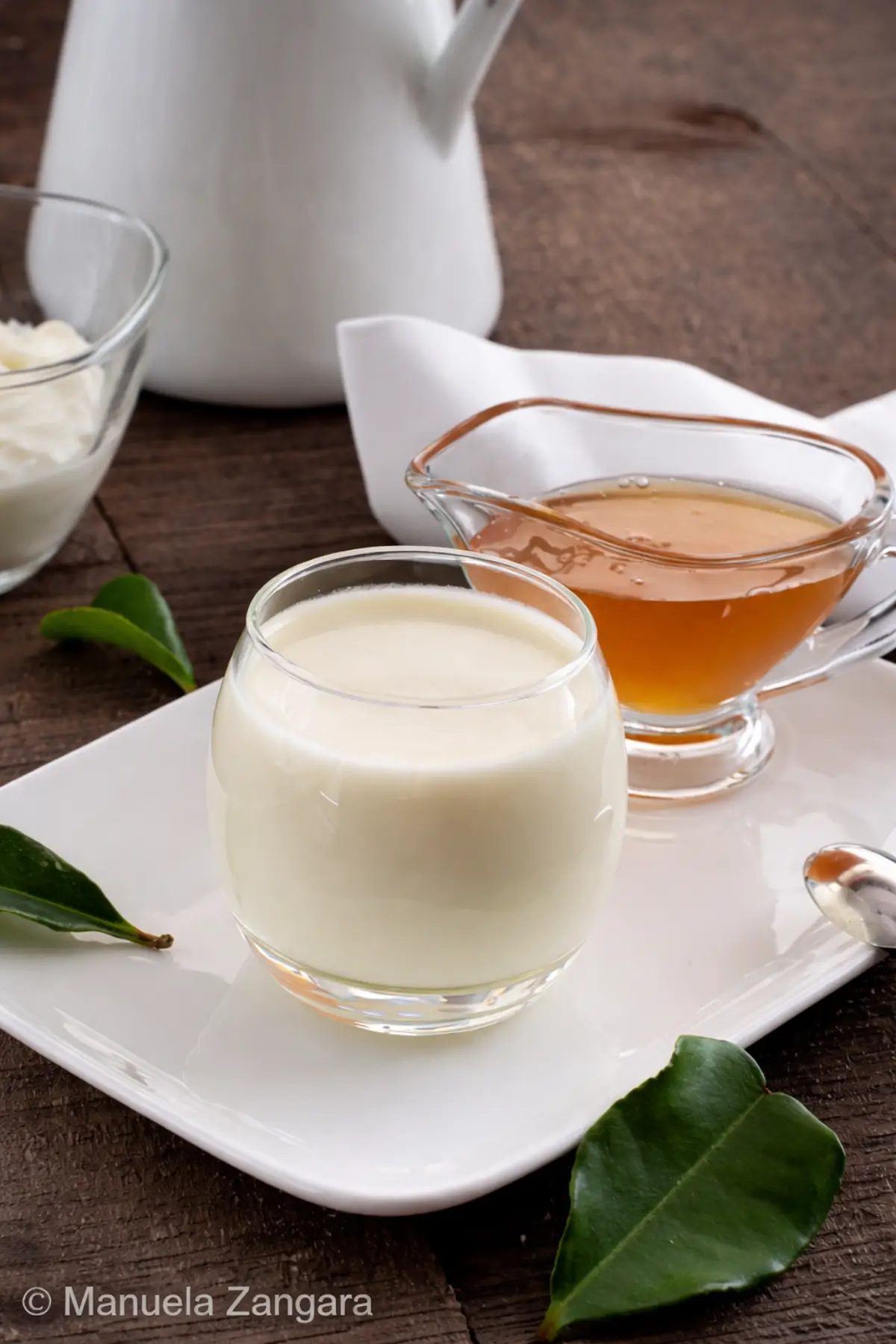 Panna cotta in a glass served with honey in a small jug on a white plate.