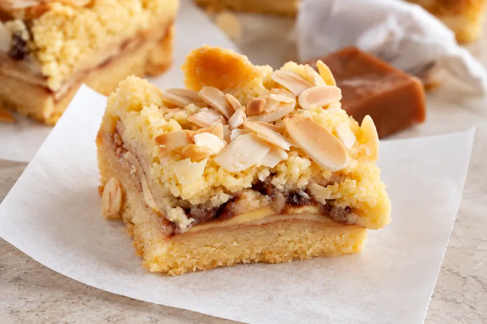 Close-up of a sliced apple bar showing its golden pastry base, apple and caramel filling, and crumbly almond topping.