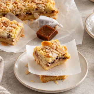 Caramel Apple Bars with layers of sliced apples, golden crumble and flaked almonds, served on white plates with wrapped caramels nearby.