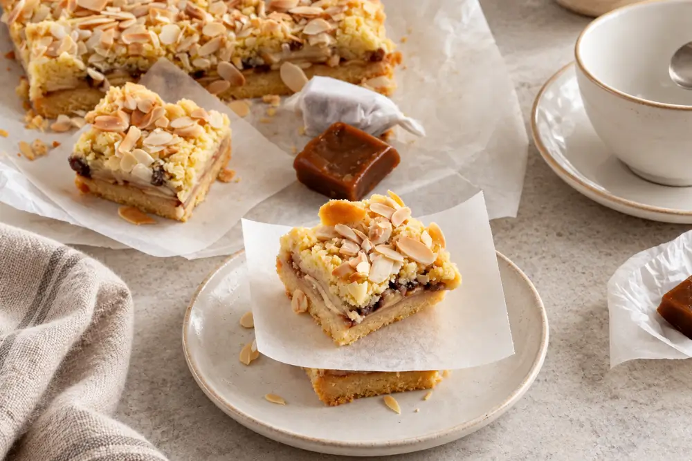 Caramel Apple Bars with layers of sliced apples, golden crumble and flaked almonds, served on white plates with wrapped caramels nearby.