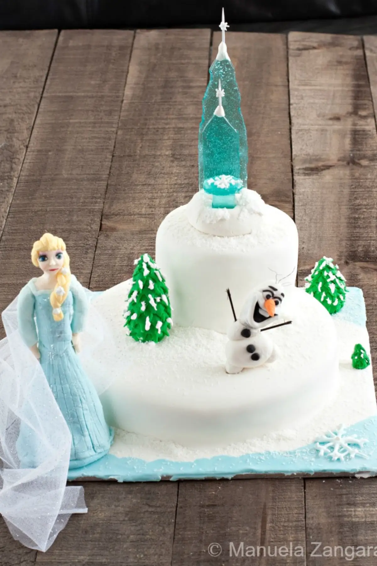 Frozen-themed two-tier cake decorated with fondant Elsa, Olaf, green trees, and a tall blue sugar castle on a snowy base.