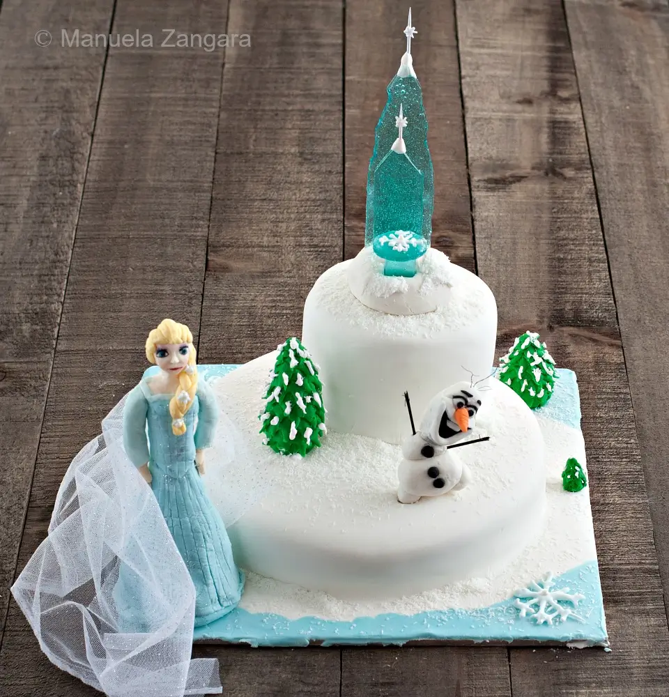 Finished Frozen-themed cake with Elsa, Olaf, sugar castle, and fondant trees on a white snowy base.