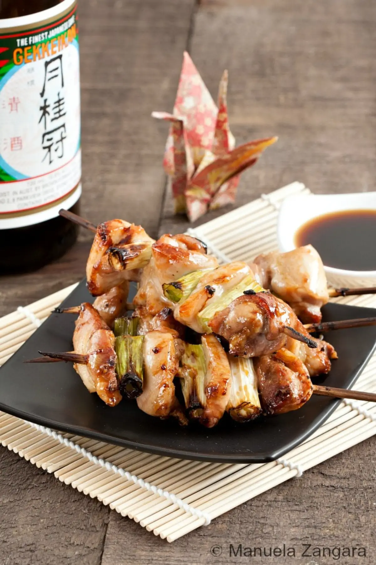 Grilled chicken skewers with spring onions glazed in a soy-based sauce, served on a black plate.