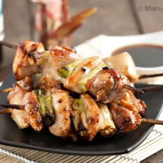 Finished yakitori chicken skewers with spring onions served on a black plate with dipping sauce.