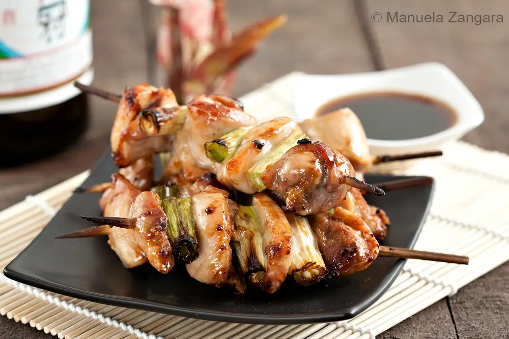 Finished yakitori chicken skewers with spring onions served on a black plate with dipping sauce.