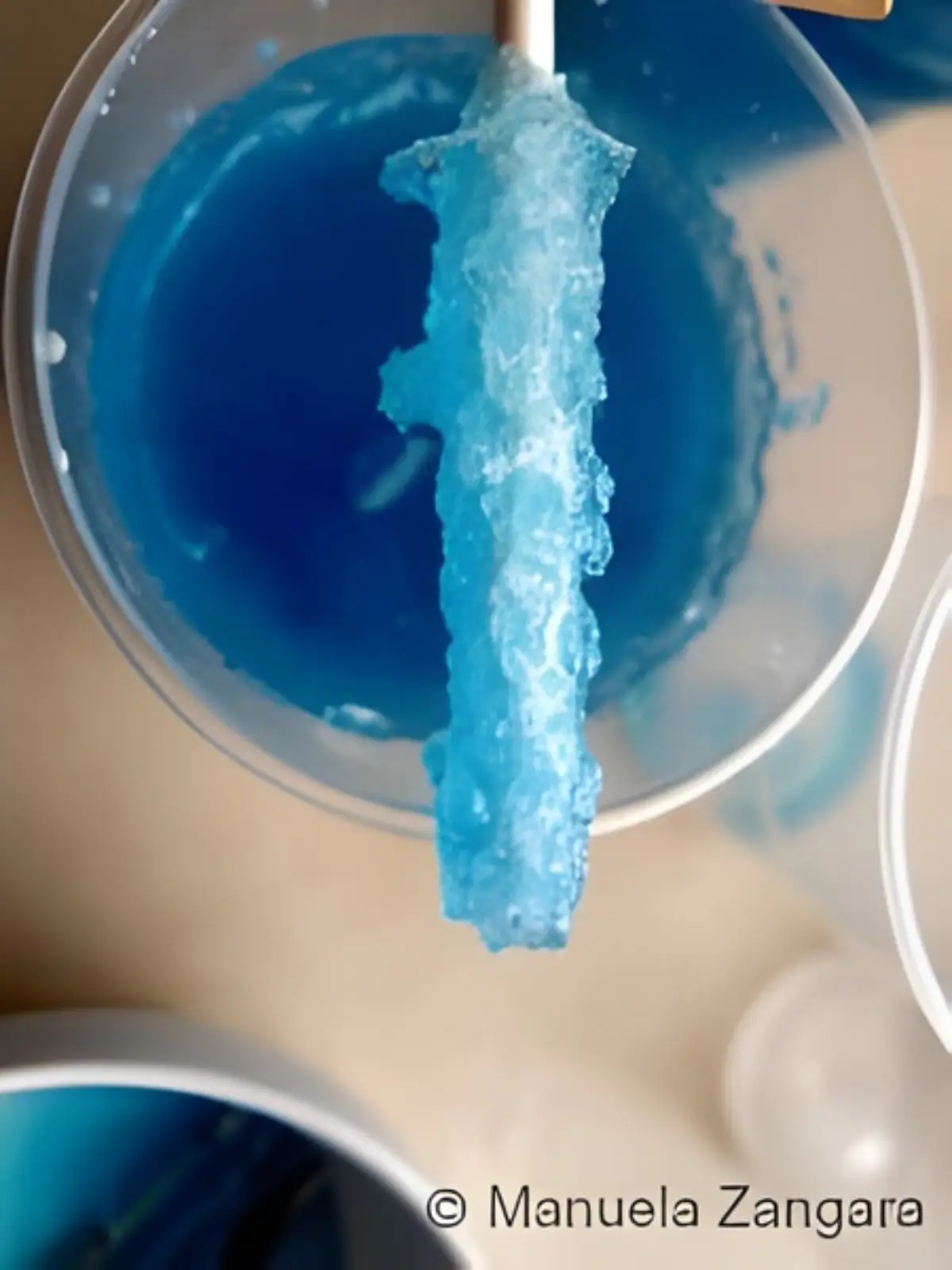 Blue rock candy lollipop forming crystals while hanging in a cup of coloured syrup.