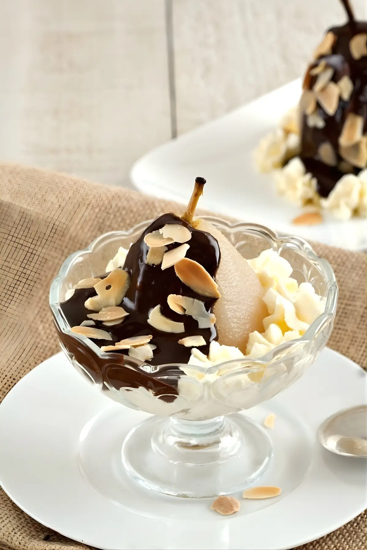 A poached pear topped with warm chocolate sauce and toasted almonds, served with whipped cream in a glass dessert bowl.