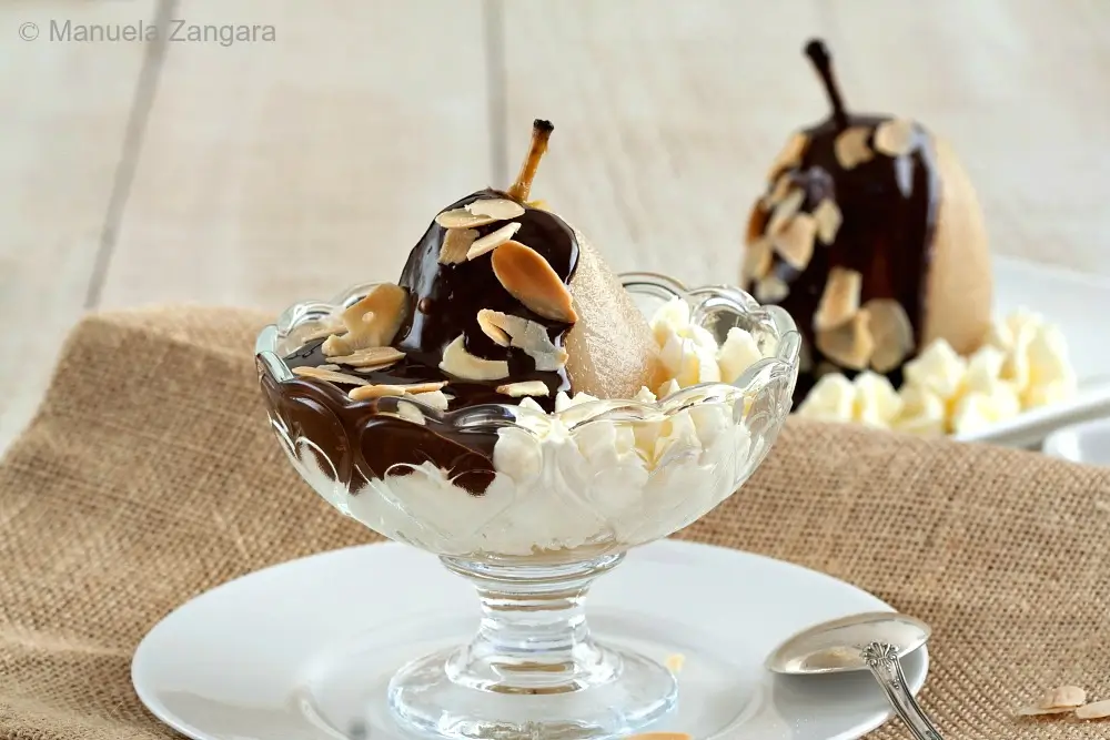 A poached pear covered in warm chocolate sauce and toasted almonds, served with whipped cream in a glass dessert dish.