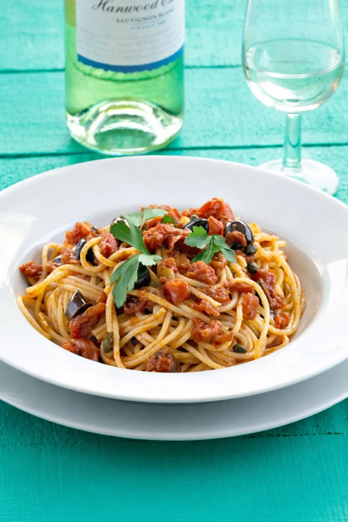 Bowl of tomato and olive pasta on a table with a glass of white wine.