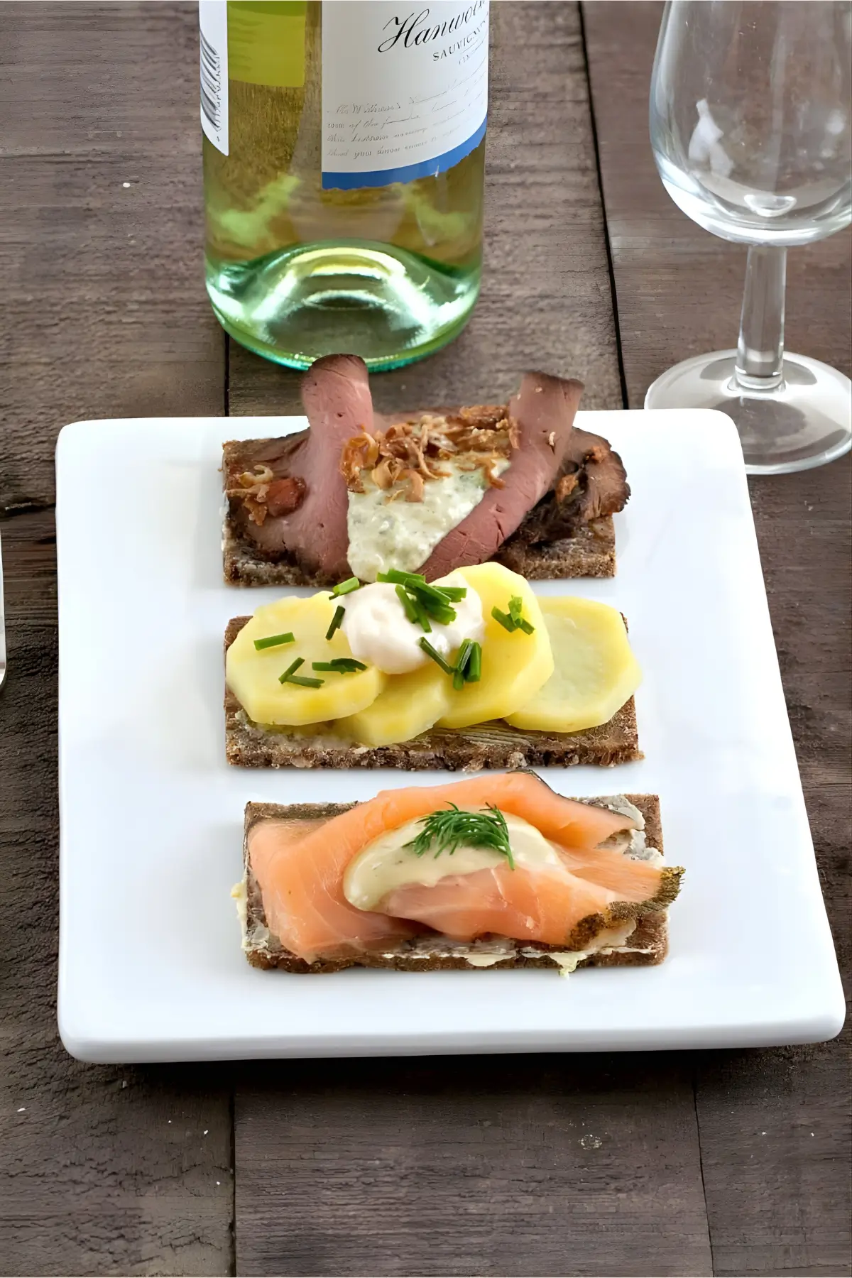 Three Scandinavian open sandwiches topped with roast beef, potatoes, and smoked salmon, served on rye bread with a glass of white wine.