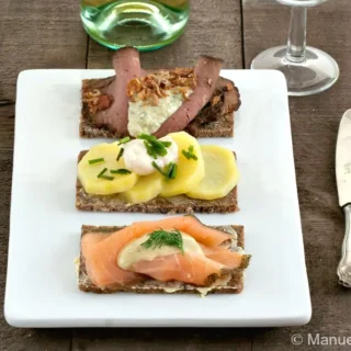 Smørrebrød platter with roast beef, potato, and salmon served on a white plate with cutlery and wine glass.