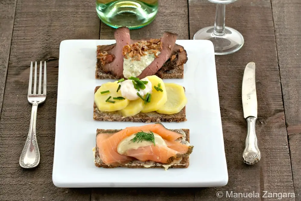 Smørrebrød platter with roast beef, potato, and salmon served on a white plate with cutlery and wine glass.