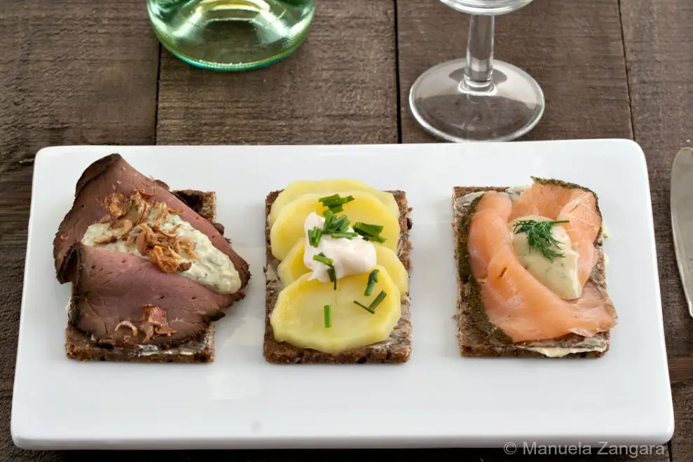 Overhead view of three Smørrebrød varieties with roast beef, potatoes, and salmon on rye bread.