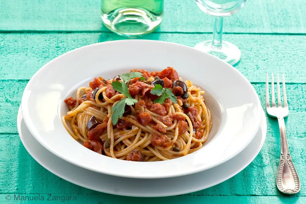 Spaghetti alla Puttanesca served in a white bowl with parsley, olives and capers.