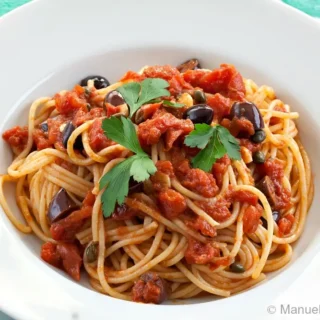 Spaghetti coated in tomato sauce with olives and capers in a white pasta bowl.