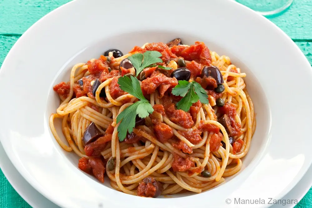 Spaghetti coated in tomato sauce with olives and capers in a white pasta bowl.