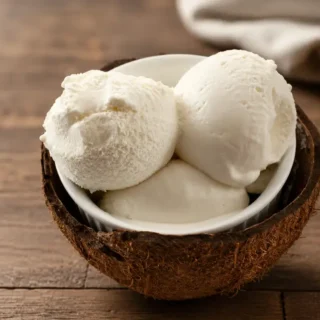 Three scoops of coconut ice cream served in a coconut shell bowl on a wooden table.