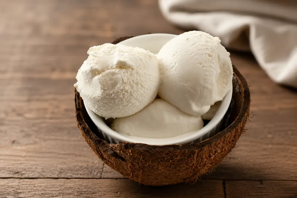 Three scoops of coconut ice cream served in a coconut shell bowl on a wooden table.