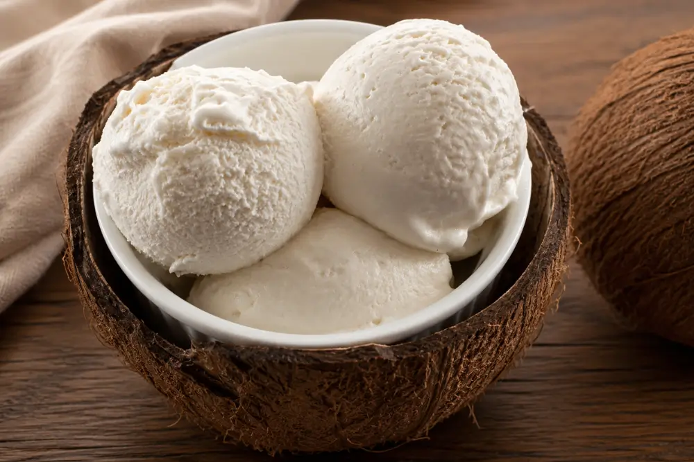Coconut ice cream served in a ramekin inside a coconut shell on a rustic wooden table.