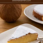 Icing sugar-dusted cake slice served with a whole coconut and remaining cake in the background.