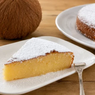 Icing sugar-dusted cake slice served with a whole coconut and remaining cake in the background.