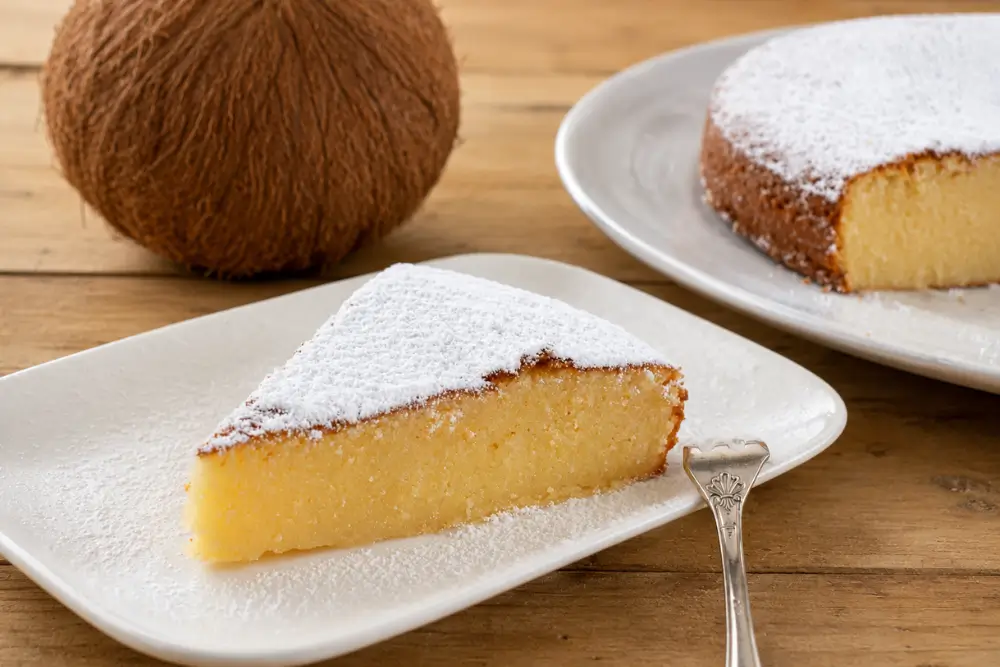 Icing sugar-dusted cake slice served with a whole coconut and remaining cake in the background.