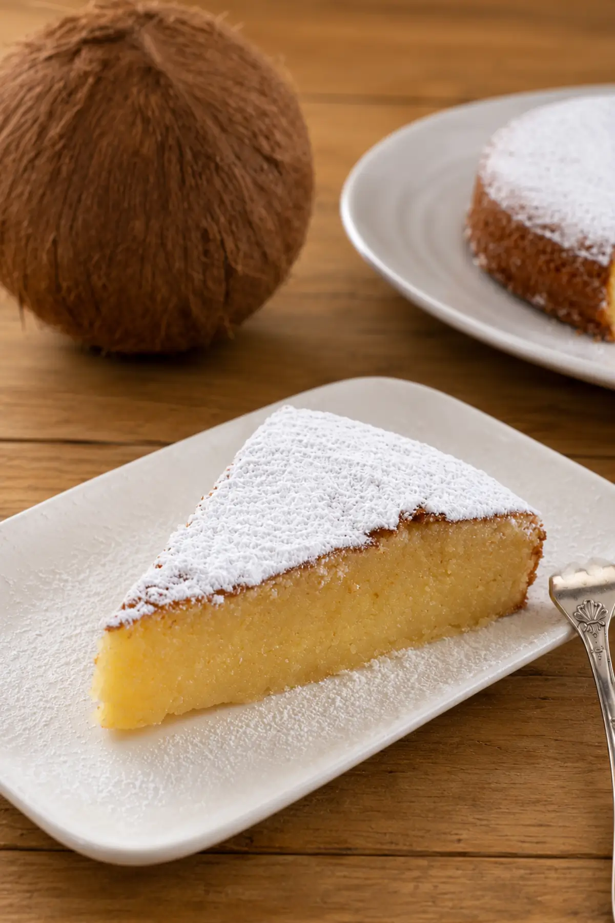 Slice of moist coconut cake dusted with icing sugar on a white plate.