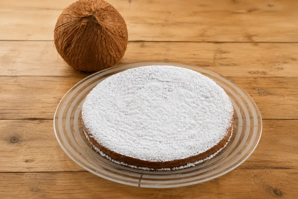 Icing sugar-dusted coconut cake on a glass serving plate with a whole coconut behind it.