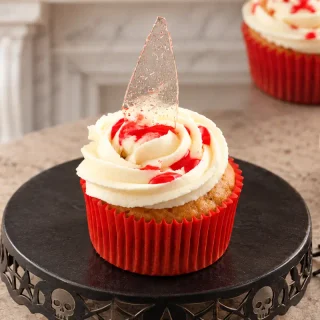 Close-up of a pumpkin cupcake topped with ivory buttercream, strawberry syrup and a pointed sugar glass shard.
