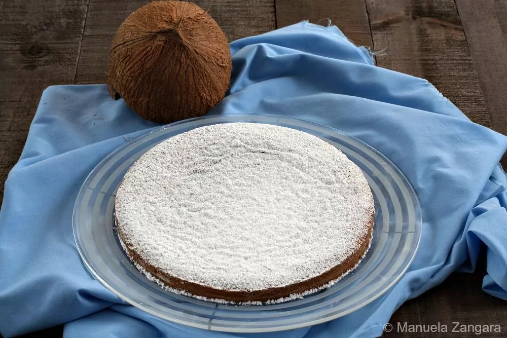 Whole condensed milk and coconut cake dusted with icing sugar, placed on a glass plate with a coconut beside it.