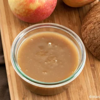 Jar of coconut caramel sauce on a wooden board with apples and coconut in the background.