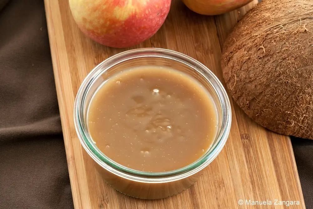 Jar of coconut caramel sauce on a wooden board with apples and coconut in the background.