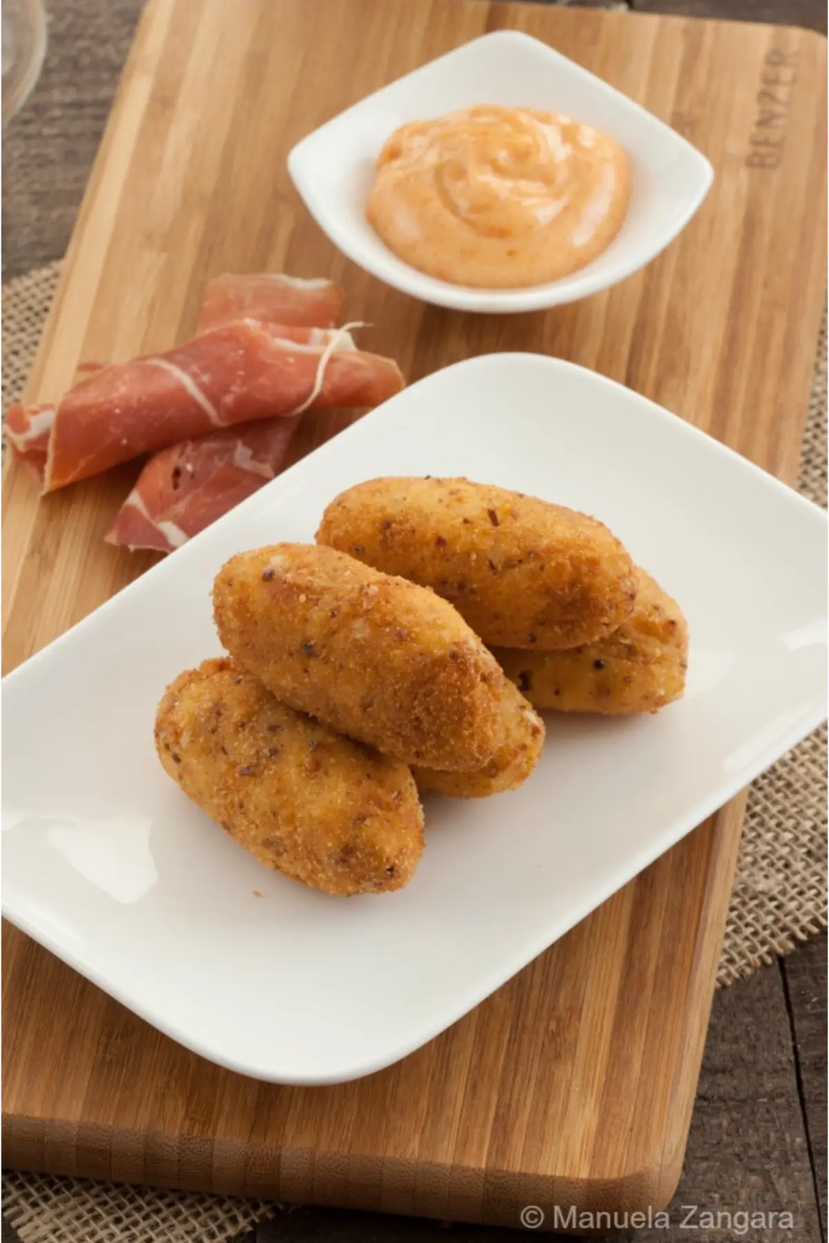 Plate of golden Serrano croquettes served with paprika mayo and slices of cured ham.