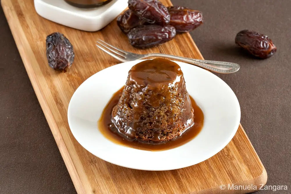 Sticky date pudding served on a white plate with dates on a wooden board.