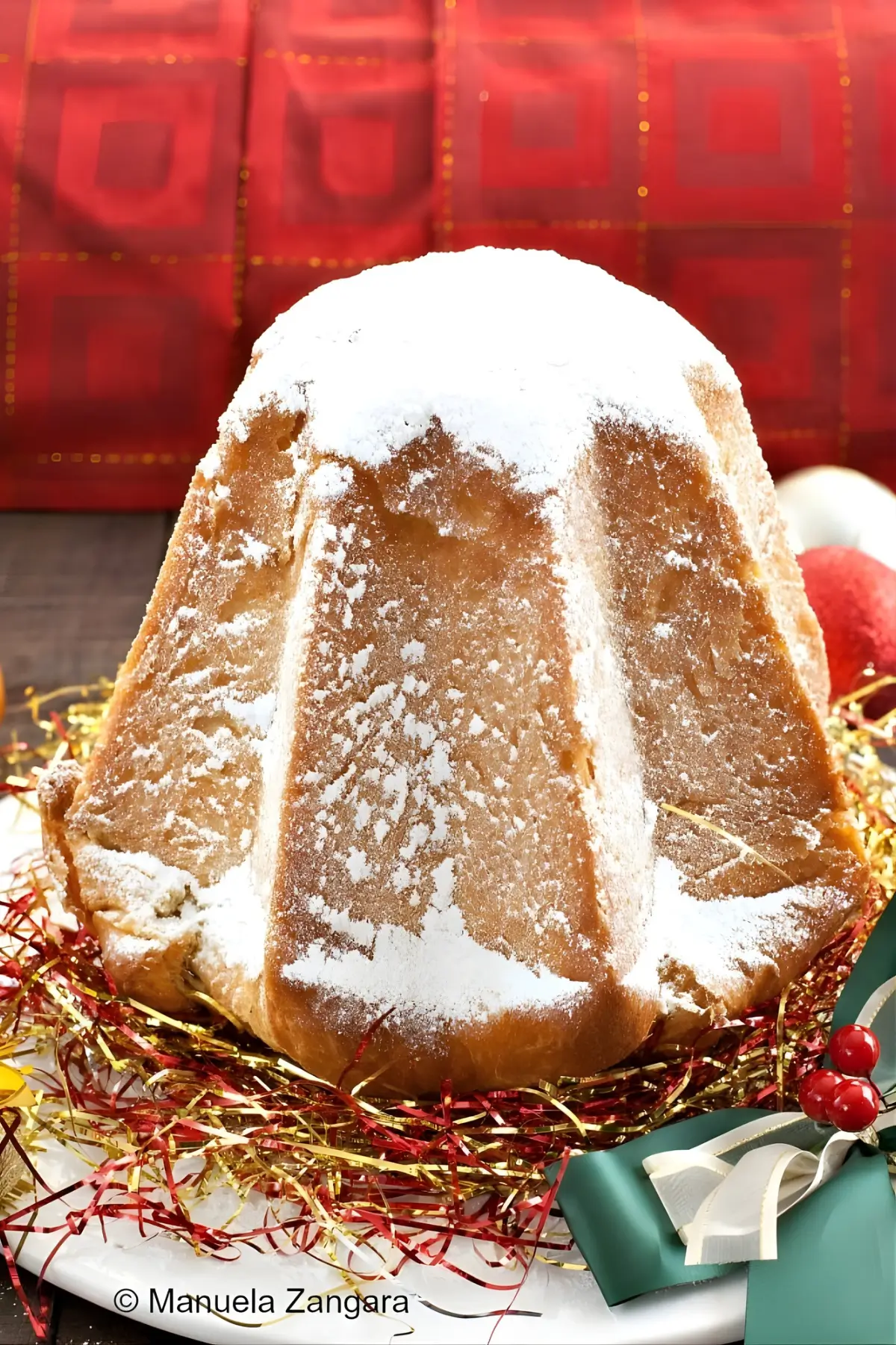Whole Pandoro dusted with icing sugar and set on a festive plate decorated with ribbons and tinsel against a red background.