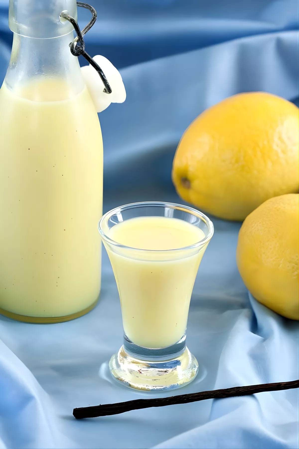 Creamy Crema di Limoncello poured into a shot glass with lemons and a vanilla pod on a blue background.