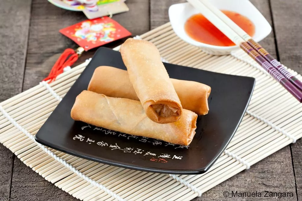 Crispy chicken spring rolls served on a black plate with chopsticks and sweet chili sauce.