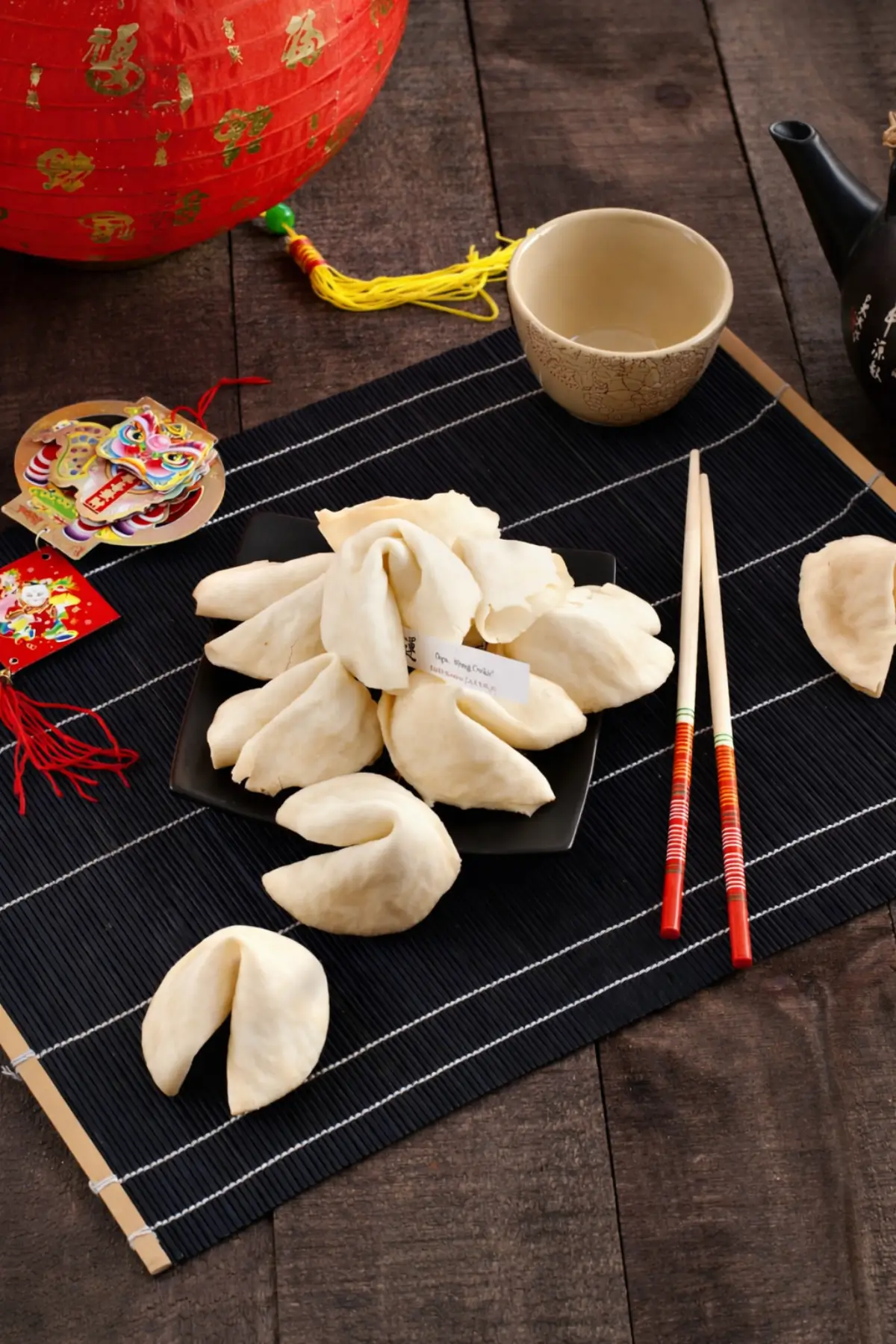 Plate of fortune cookies arranged on a black dish with chopsticks and tea set.
