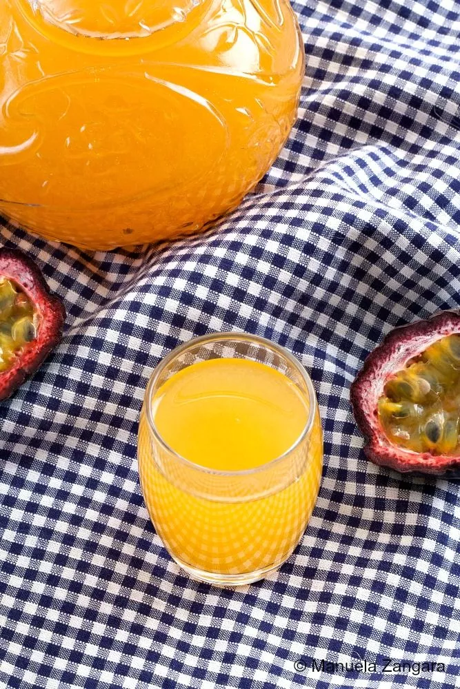 Glass of golden homemade passionfruit liqueur served on a blue gingham cloth with fresh passion fruit.