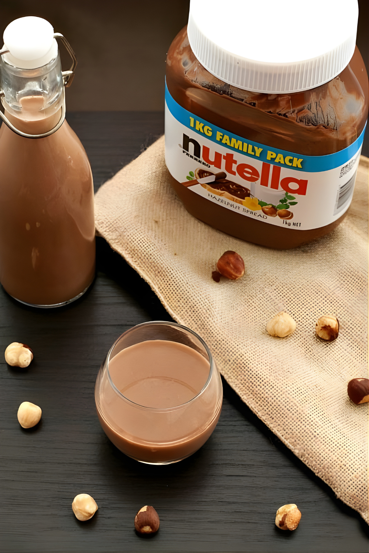 Homemade Nutella Liqueur in a small glass and bottle with hazelnuts and a large Nutella jar in the background.