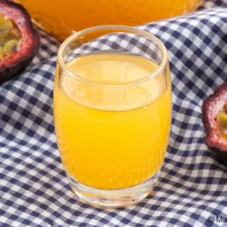 Glass of homemade Passion Fruit Liqueur on a checkered cloth with fresh passion fruit halves beside it.