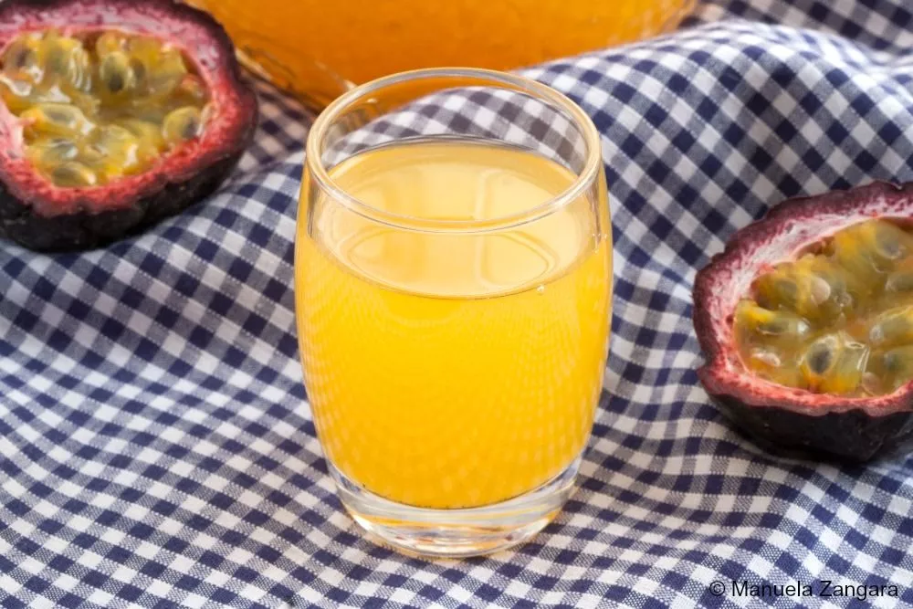 Glass of homemade Passion Fruit Liqueur on a checkered cloth with fresh passion fruit halves beside it.