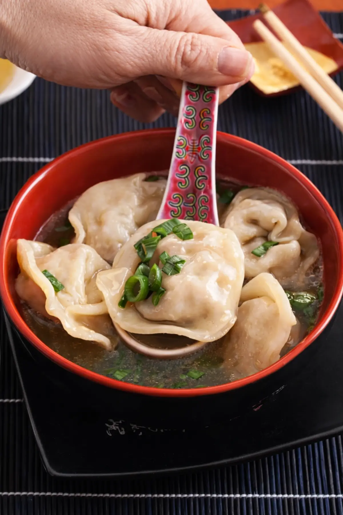 Spoon lifting wontons from a bowl of wonton soup with green onions.