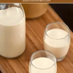 Creamy Dulce de Leche Liqueur served in small glasses on a wooden board beside a spoonful of caramel.