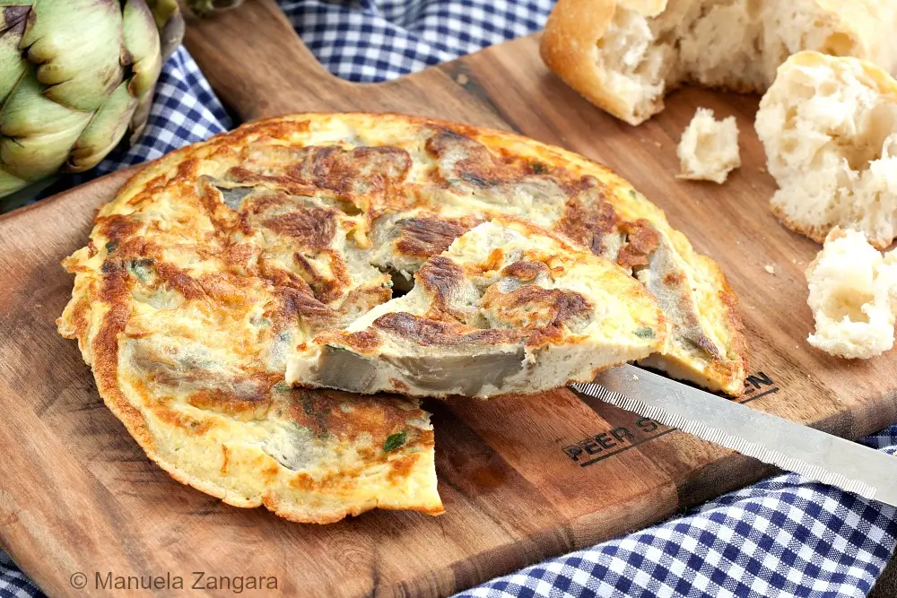 Cooked artichoke frittata sliced on a serving plate with bread and fresh artichokes in background.