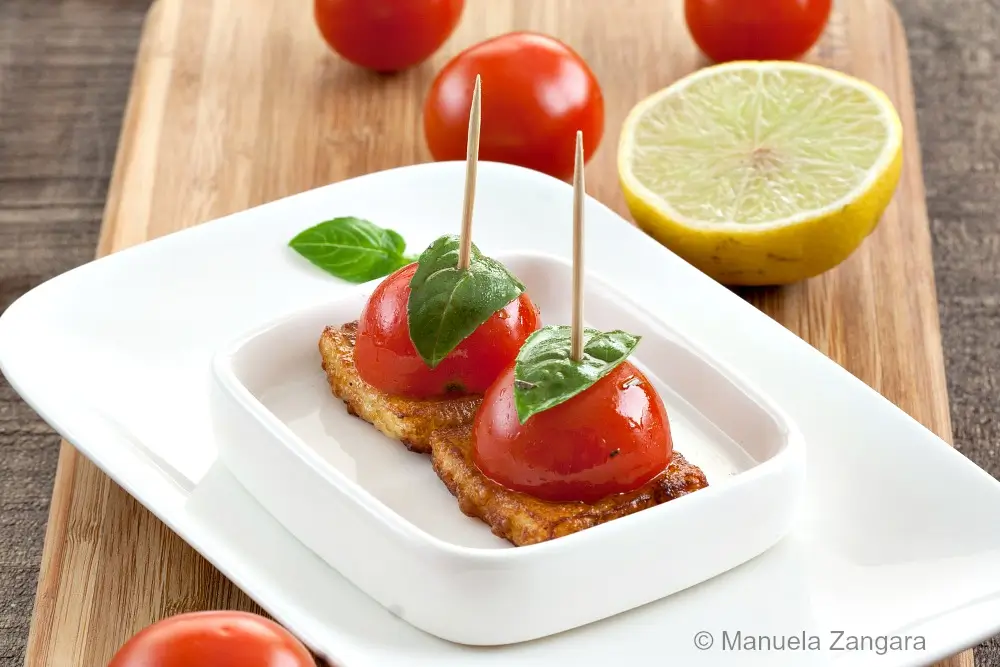Cherry tomato and basil skewers placed on fried halloumi squares, served on a white dish with lemon and tomatoes in the background.