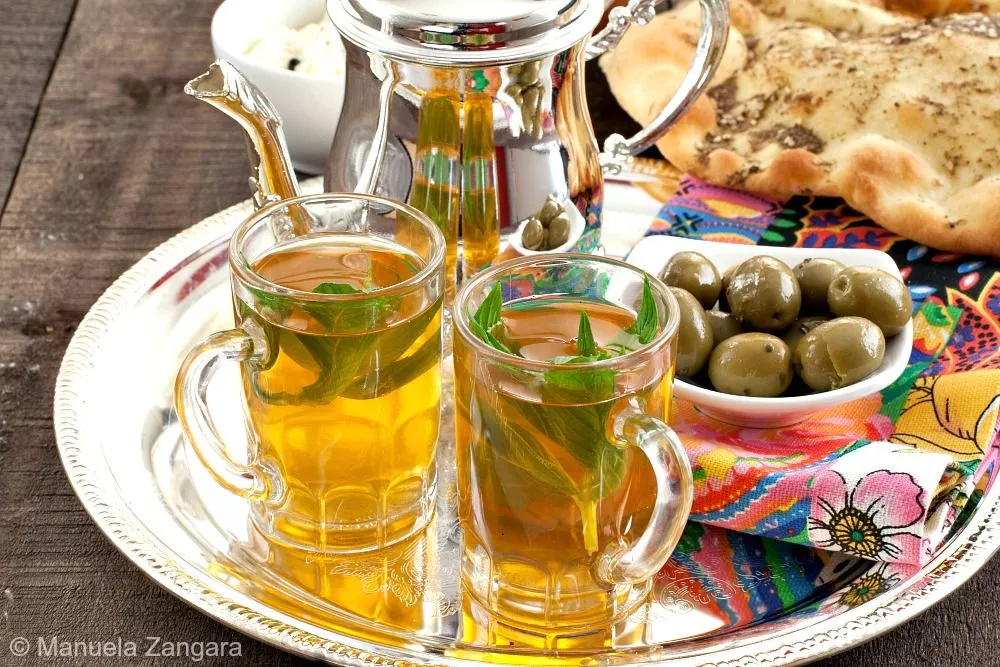 Moroccan mint tea served in glasses with flatbread, olives, and cheese on a colourful tray.